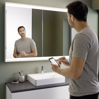 Un homme commande lʼéclairage de lʼarmoire à miroir Geberit ONE avec son smartphone (© Geberit)